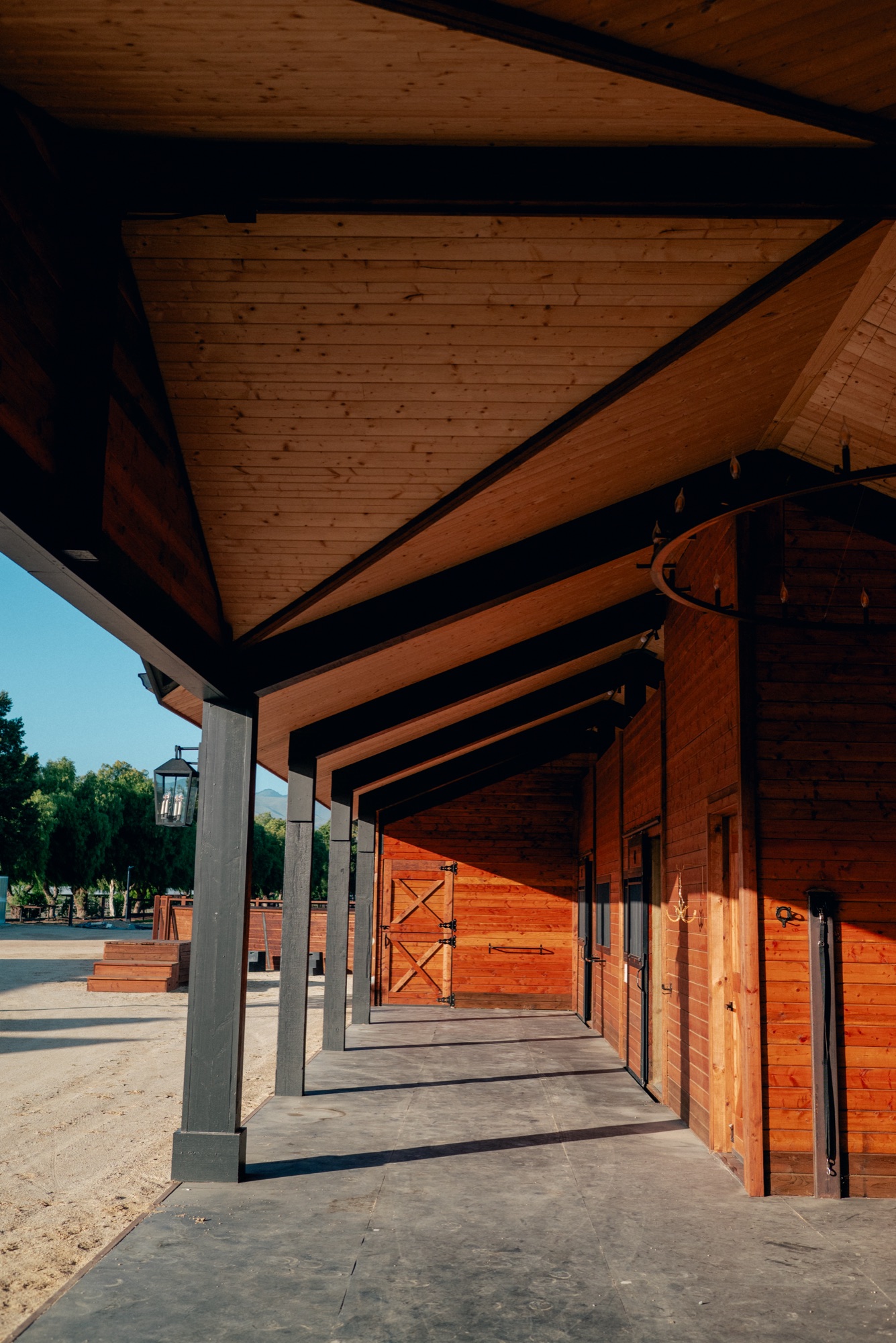 The shaded stable breezeway at golden hour, stall doors in warm redwood