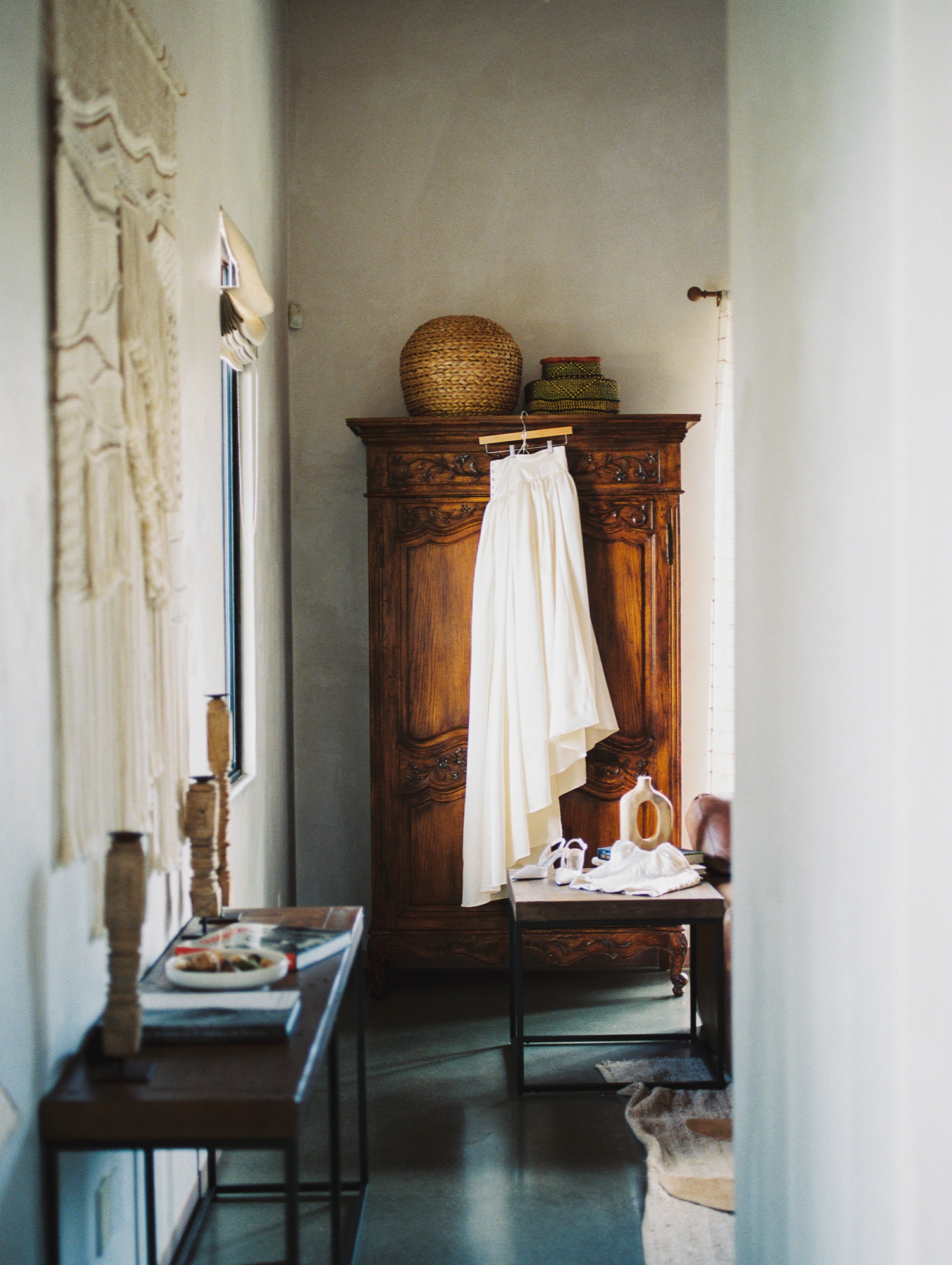 A wedding gown hanging ready beside a carved armoire in the getting-ready suite